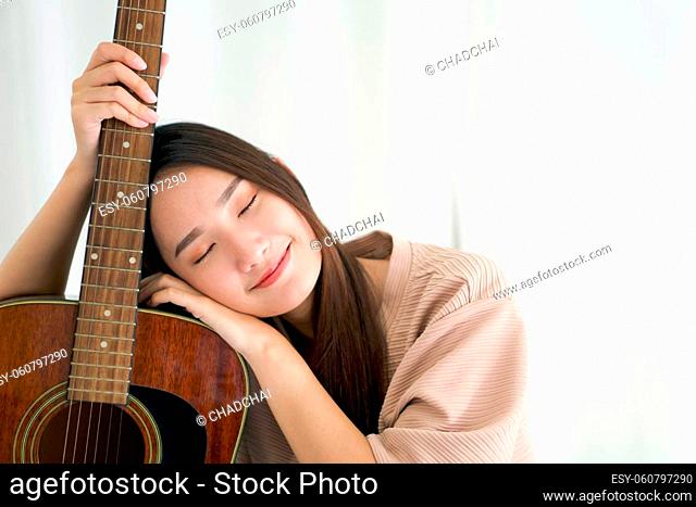 Asian woman in brown T-shirt with eyes closed smiling happily leaning on guitar. After completing the practice of playing music, enjoy leisure weekend at home