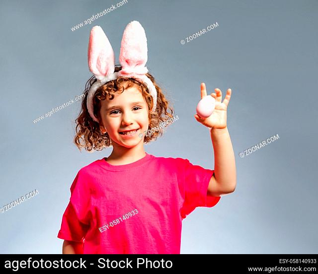 The smiling little girl with bunny ears holding an easter egg. The girl with curly hair stands on a gray background