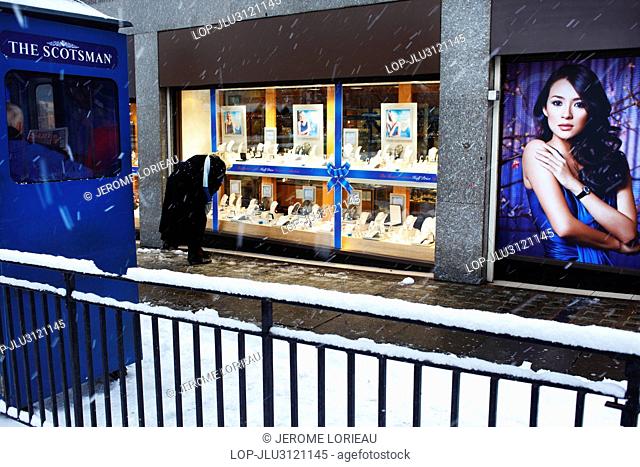 Scotland, City of Edinburgh, Edinburgh. A woman looking in the window display of a jewellery shop opposite a newspaper stand whilst the snow falls on Princes...