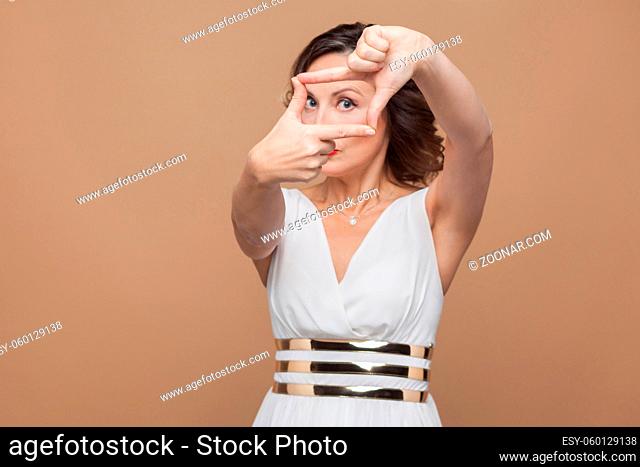 Imagination look. Woman showing frame sign at camera. Emotional expressing woman in white dress, red lips and dark curly hairstyle