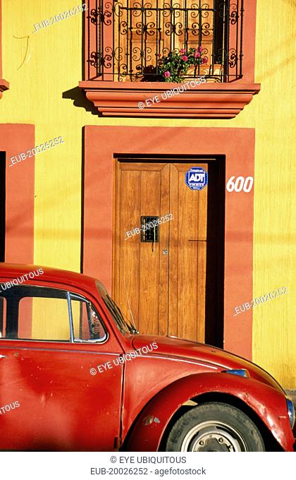 Partly seen red volkswagon beetle outside yellow building with wooden door and window shutters in orange painted frames