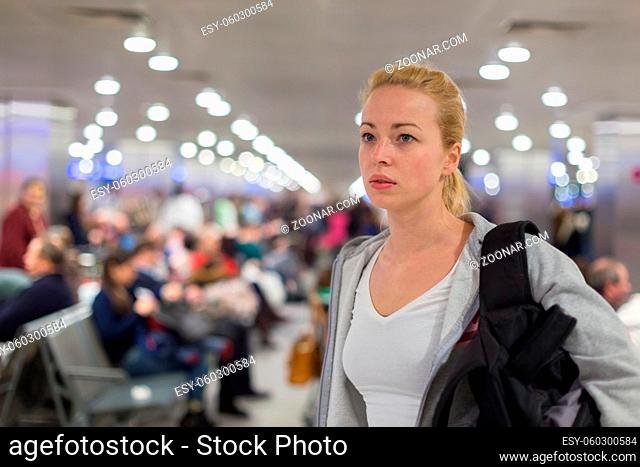 Young blond caucsian woman waiting on airport terminal full of passenegers waiting to depart