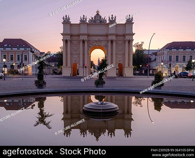 28 September 2023, Brandenburg, Potsdam: Behind the Brandenburg Gate in Potsdam, the sky turns colorful at sunrise and is reflected in the water of the fountain...