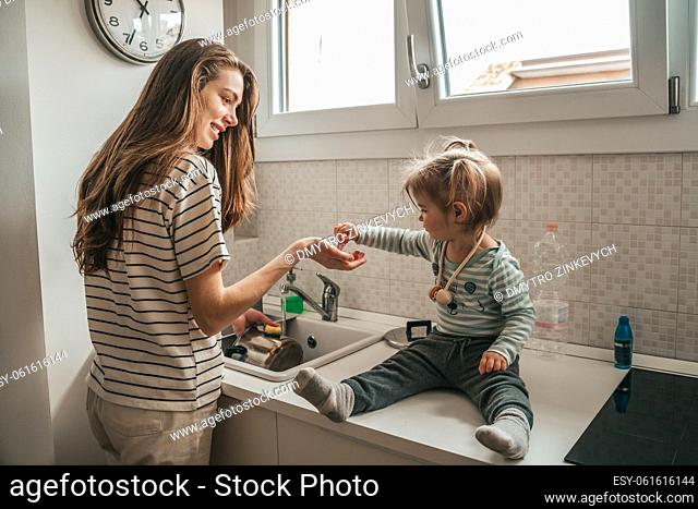 Little girl sitting on the kitchen sink helping her happy young mother to wash the cooking pot