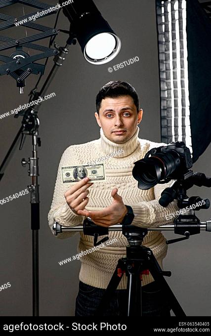 Professional handsome photographer with digital camera on tripod showing hundred dollar bill on gray background with lighting equipment