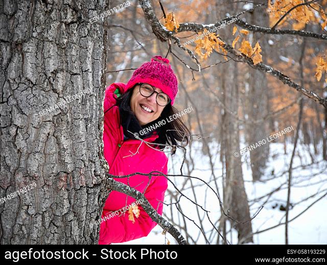 In the snowy forest, a woman dressed in mountain clothes, a pink down jacket, gloves and a wool hat, smiles happily in nature