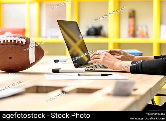 Female is using a laptop on wooden table on the blurry background of the yellow shelves in the office. Next to her there are football ball, markers and pencils