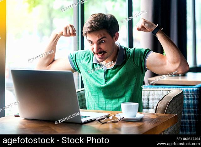 I am strong. Young aggressive businessman in green t-shirt sitting, looking at laptop screen on video call, showing his biceps muscle, angry face