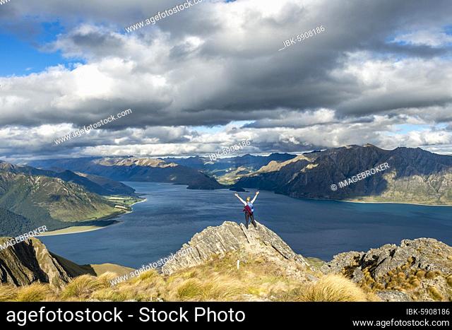 Hiker stands on a rock and stretches her arms in the air, view over Lake Hawea, lake and mountain landscape in the evening light, view from Isthmus Peak, Wanaka