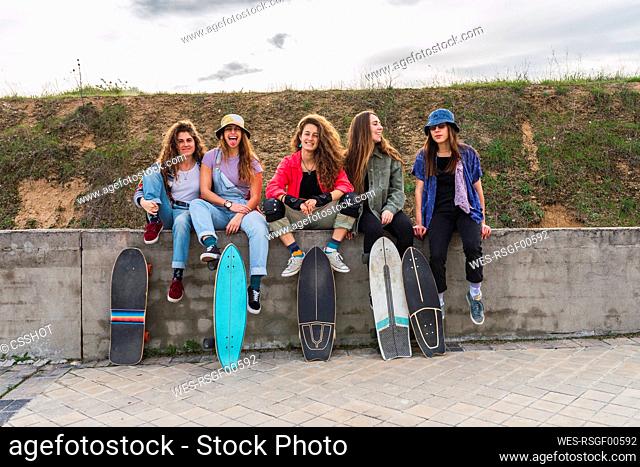 Cheerful female friends with skateboards sitting on retaining wall