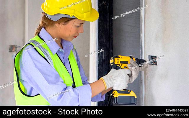 Construction worker in green reflective safety vest and hardhat use a yellow cordless electric drill with battery to drill holes in the wall socket
