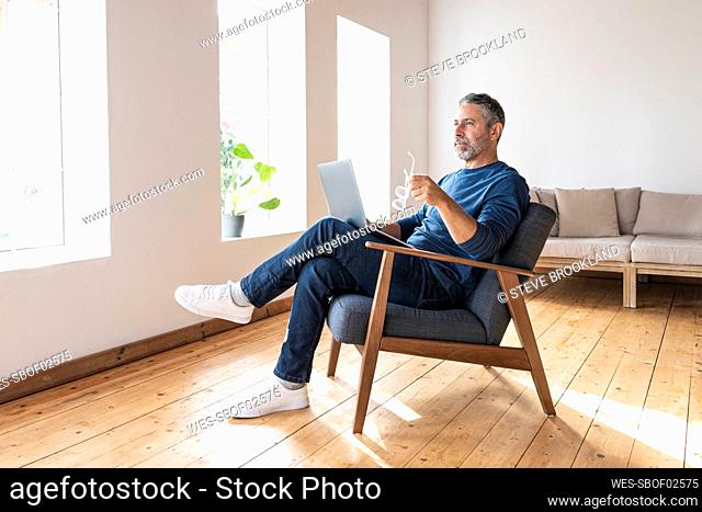 Thoughtful businessman holding eyeglasses while sitting with laptop at home