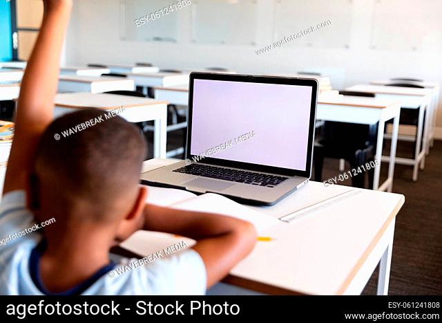 African american elementary schoolboy with hand raised using laptop at desk in classroom