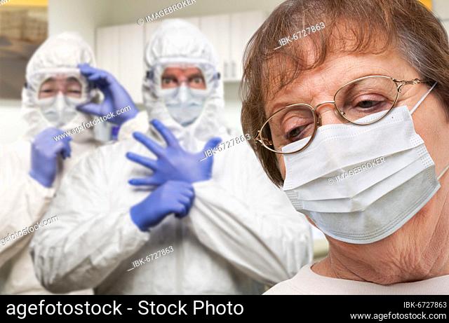 Senior adult woman wearing medical face mask in doctor office with nurses in HAZMAT suits behind