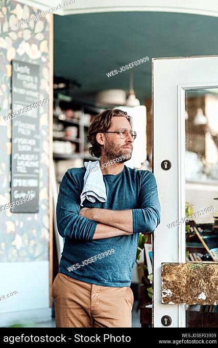 Male owner with arms crossed leaning at door in coffee shop