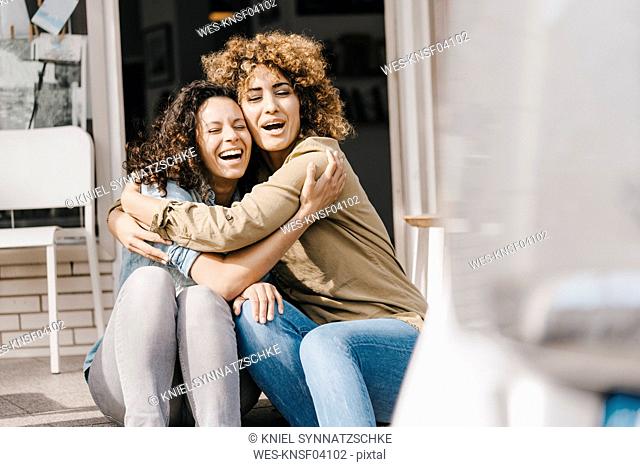 Two laughing friends sitting in front of coworking space, embracing