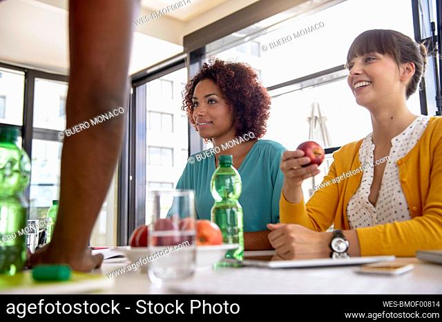 Smiling woman holding apple while sitting with female colleague in office