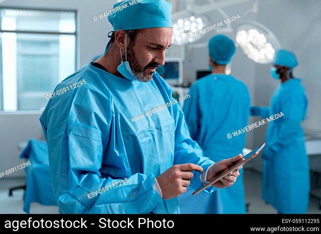 Caucasian male surgeon with face mask and protective clothing using tablet in operating theatre