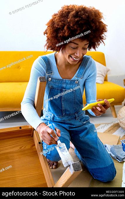 Smiling redhead woman using mobile phone while painting chair at home