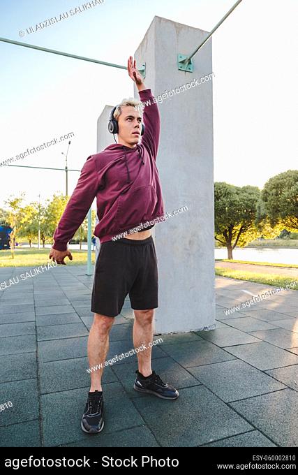 Fitness man warming up outdoors on sports ground and listen to music in headphones. Rotates body and swings hands