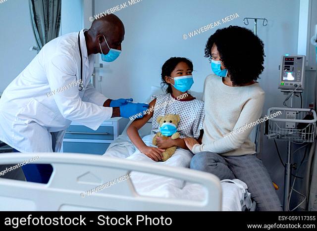 Male doctor giving covid vaccination to sick girl in hospital bed with mother, all in face masks