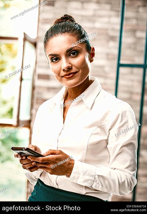 Cheerful Caucasian Woman Looks At Camera Using Phone, Young Women Typing Message By Phone Standing Near Opened Window In Office, Toned Image