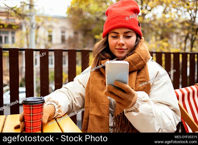 Woman wearing knit hat and scarf using smart phone while sitting at sidewalk cafe