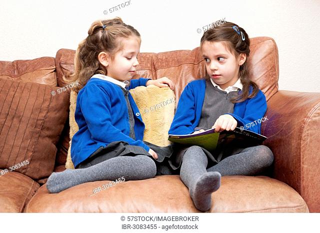Young sisters in Primary School uniform reading a book together, sitting on a sofa, United Kingdom
