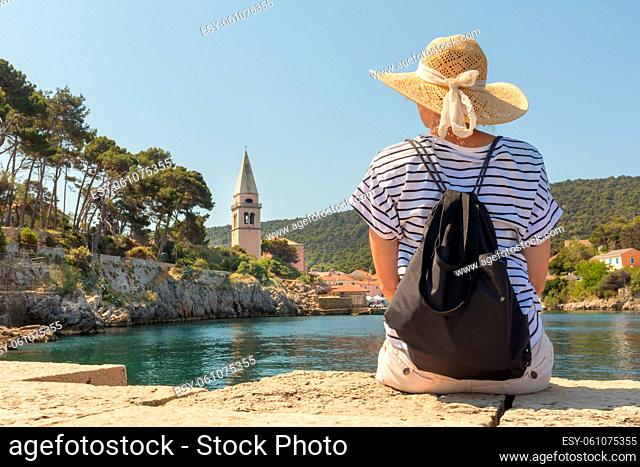 Woman traveler wearing straw summer hat and backpack, sittingat edge of stone pier, enjoying beautiful panoramic view of Veli Losinj, Losinj island, Croatia