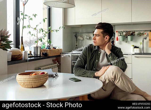 Young man looking through window sitting at table in kitchen