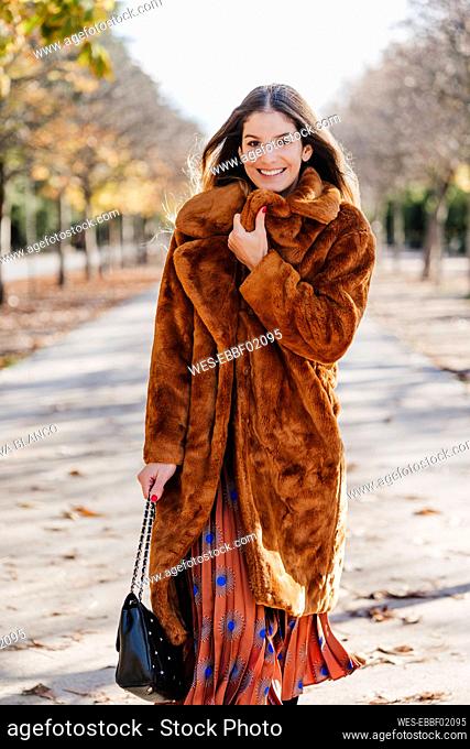 Happy woman in fur coat walking while holding purse on footpath