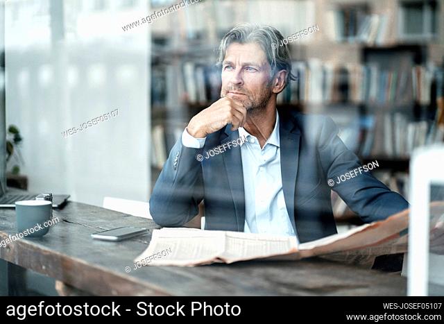 Thoughtful male professional sitting with hand on chin in cafe seen through glass window