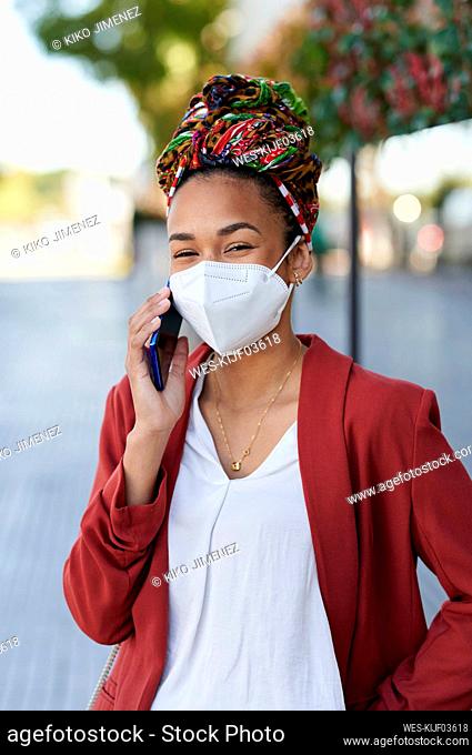 Young woman wearing headscarf and protective face mask talking on mobile phone while standing outdoors