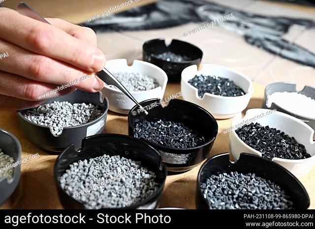 PRODUCTION - 07 November 2023, Baden-Württemberg, Pforzheim: Oliver Lemke removes an iron-on bead from bowls containing iron-on beads in various shades of gray...