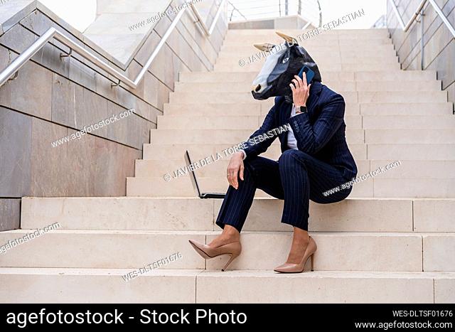 Female stockbroker with laptop wearing bull mask talking on smart phone while sitting on staircase