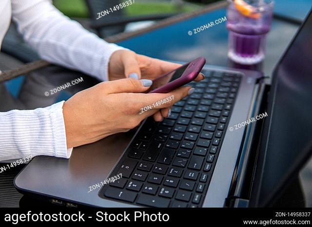Modern business woman is reading incoming sms message on smartphone or watching broadcasting online on modern mobile phone while sitting in caffe with a laptop