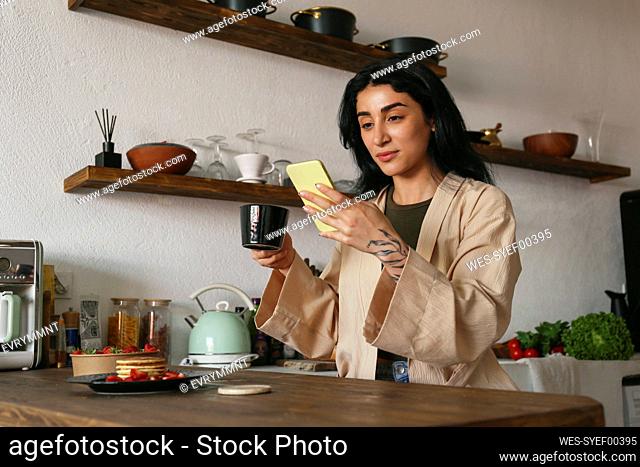 Young woman using smart phone and enjoying brewed coffee at table