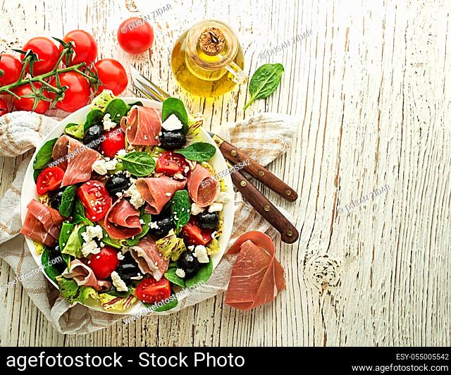 Healthy green salad with prosciutto, olives, tomato and fresh cheese on wooden table background. Healthy meal