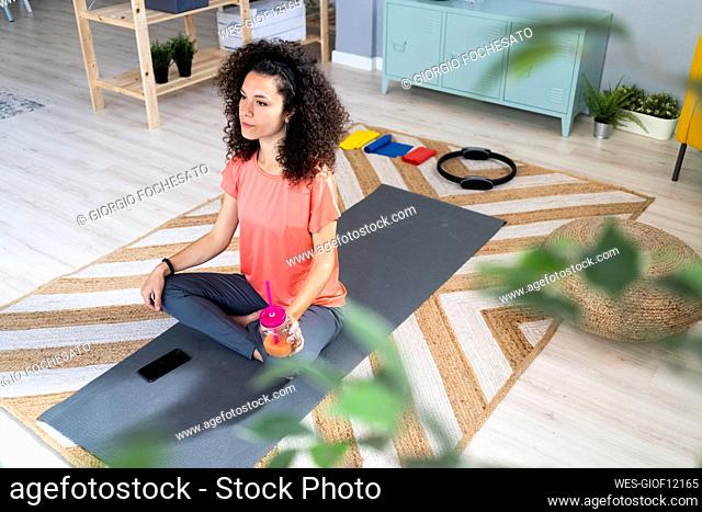 Contemplating woman holding juice jar while sitting on exercise mat in living room