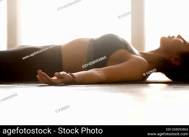 Young sporty attractive woman practicing yoga, doing Dead Body exercise, Savasana, Corpse pose, working out, wearing sportswear, grey top, indoor close up