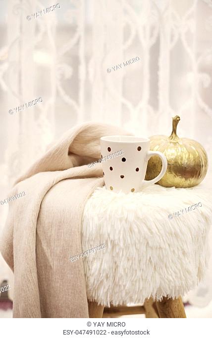 Hot chocolate, a cup of cappuccino on a fur chair in front of a large window with a white sheers curtain. Warm scarf and golden pumpkin around