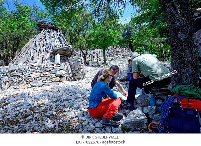 A young woman with trekking gear discussing the trail on a map with locals in front of an Ovile, which is a traditional shepherds hut, Selvaggio Blu, Sardinia