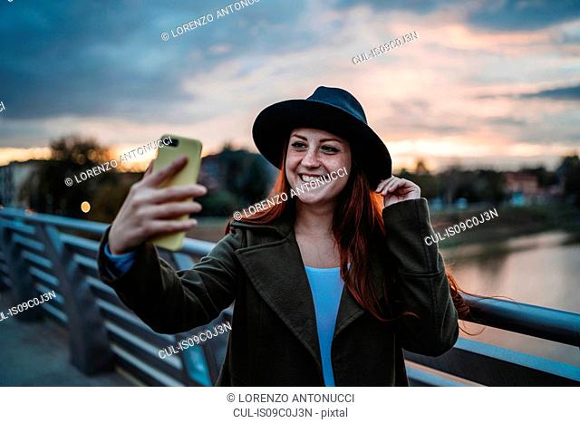 Young woman with long red hair on footbridge taking smartphone selfie at dusk, Florence, Tuscany, Italy