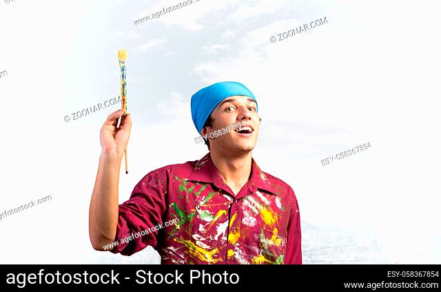 Smiling young painter artist pointing upwards with paintbrush in hand. Portrait of happy painter on modern downtown background