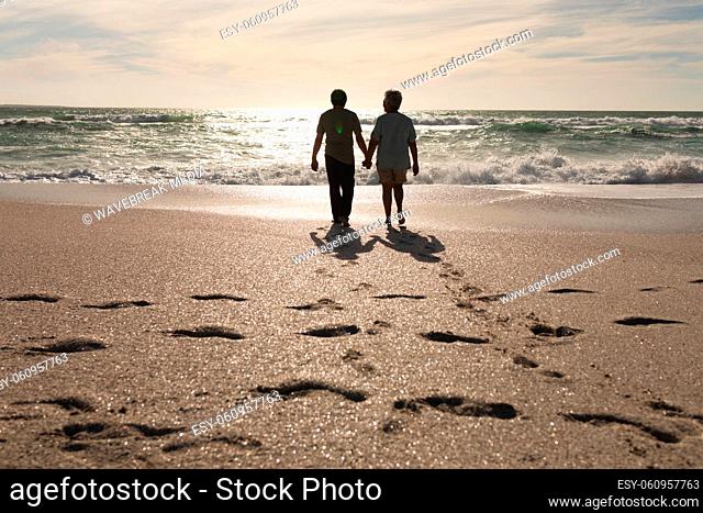 Full length of senior multiracial couple holding hands walking at beach with footprints on sunny day