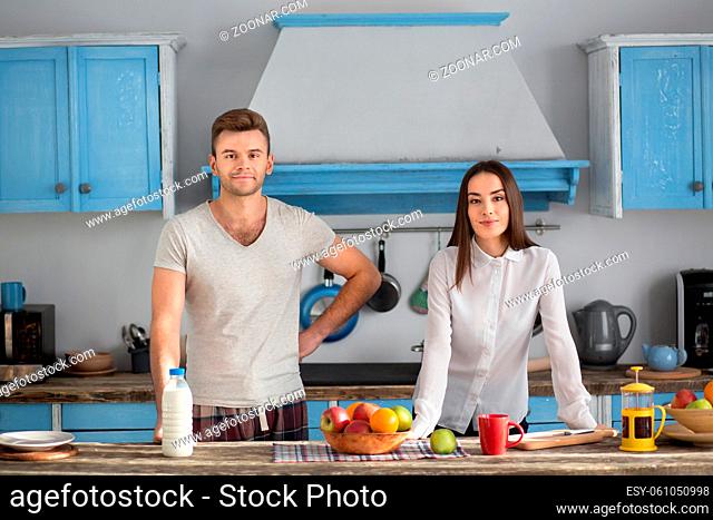 Happy young couple at kitchen in the morning. Man and woman smiling standing at table id daylight in their cuisine
