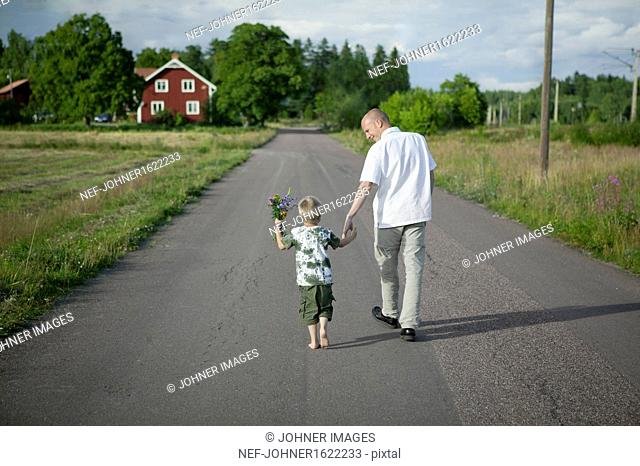 Father walking with son on road, boy holding flowers
