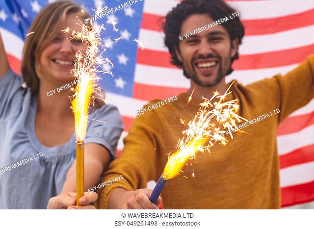 Portrait of young Caucasian couple playing with fire cracker while holding american flag. Thhey are smiling and looking at camera