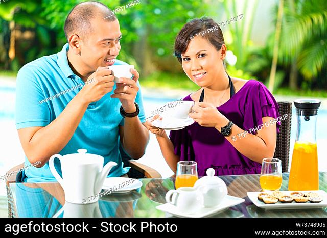 Asian couple having coffee on the porch in front of their home, in the background a tropical garden