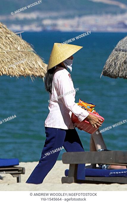 Woman beach vendor in conical hat walks between woven beach umbrellas Nha Trang south east Vietnam